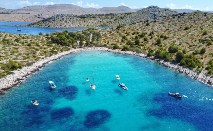 Aerial view of a turquoise bay near Dubrovnik with several boats anchored and people swimming by a rocky shoreline with sparse vegetation, perfect for private boat tours or day trips.