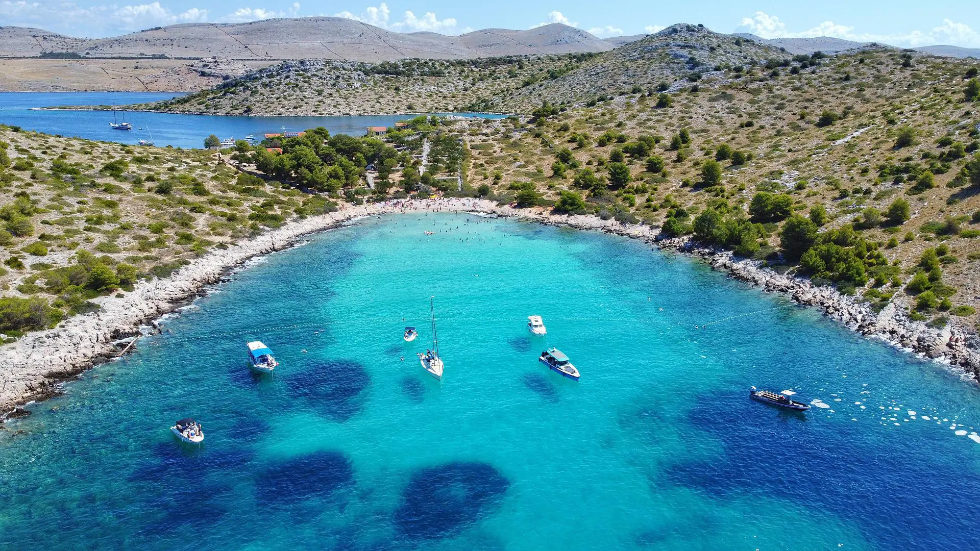 Aerial view of a turquoise bay near Dubrovnik with several boats anchored and people swimming by a rocky shoreline with sparse vegetation, perfect for private boat tours or day trips.