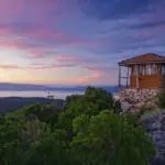 A small, wooden lookout tower sits atop a rocky hill surrounded by dense green foliage, overlooking a serene body of water at sunset with a sky painted in shades of pink and blue—perfect for one of those peaceful Dubrovnik day trips.