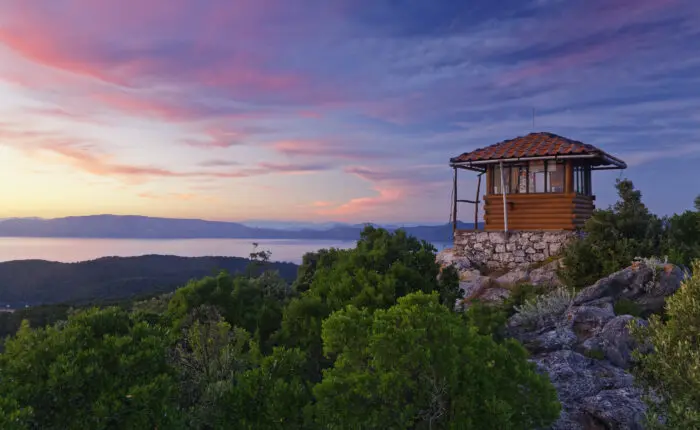 A small, wooden lookout tower sits atop a rocky hill surrounded by dense green foliage, overlooking a serene body of water at sunset with a sky painted in shades of pink and blue—perfect for one of those peaceful Dubrovnik day trips.