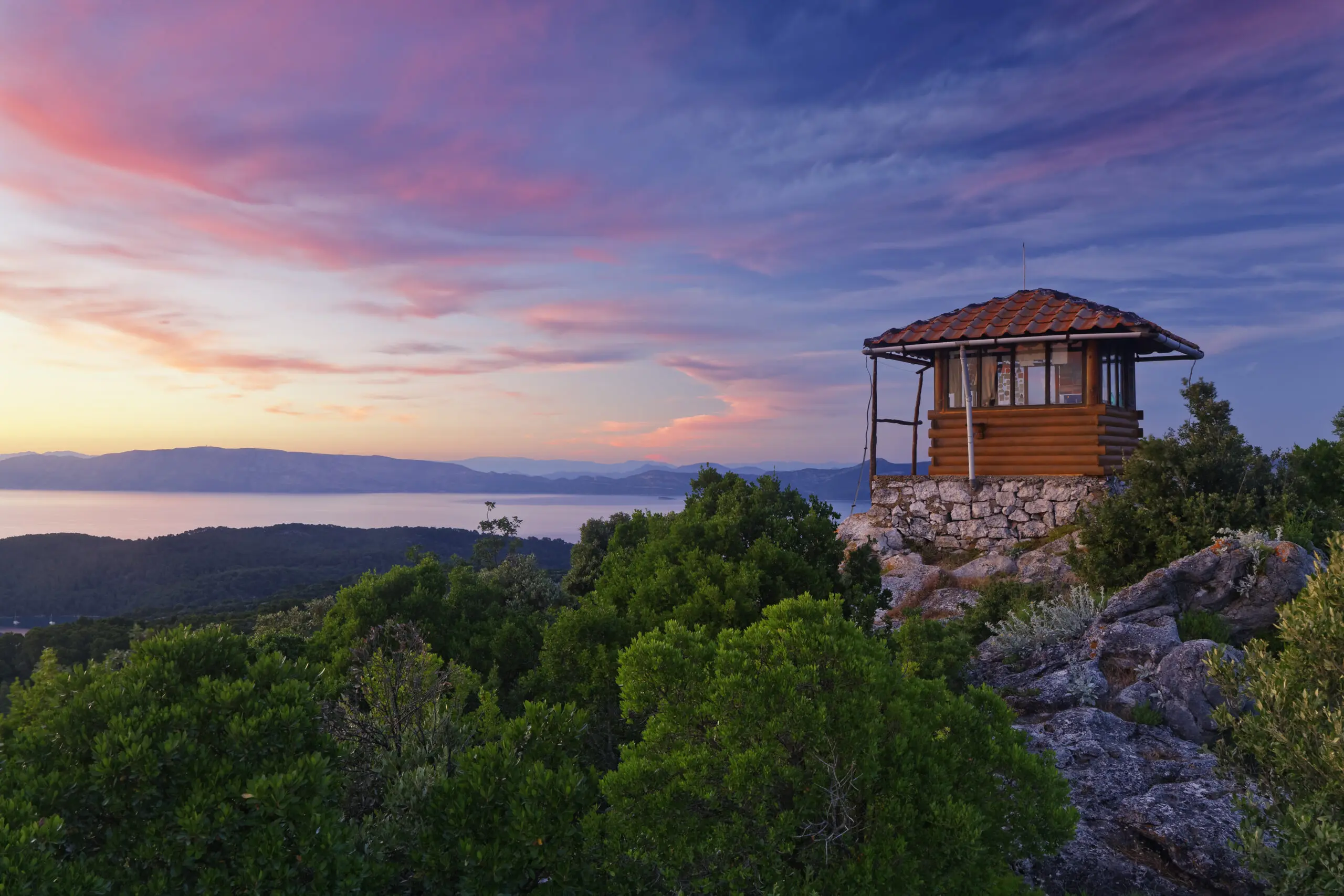 A small, wooden lookout tower sits atop a rocky hill surrounded by dense green foliage, overlooking a serene body of water at sunset with a sky painted in shades of pink and blue—perfect for one of those peaceful Dubrovnik day trips.