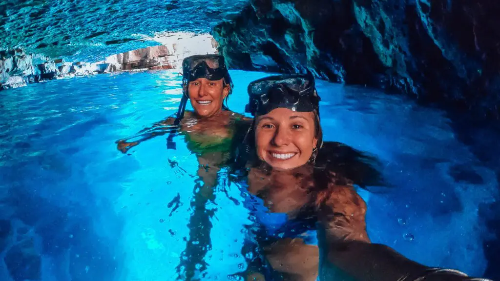 Two people wearing snorkeling gear smile in an illuminated blue cave pool, partially submerged in the vibrant water with rocky walls in the background, enjoying one of Dubrovnik's exclusive private boat tours.