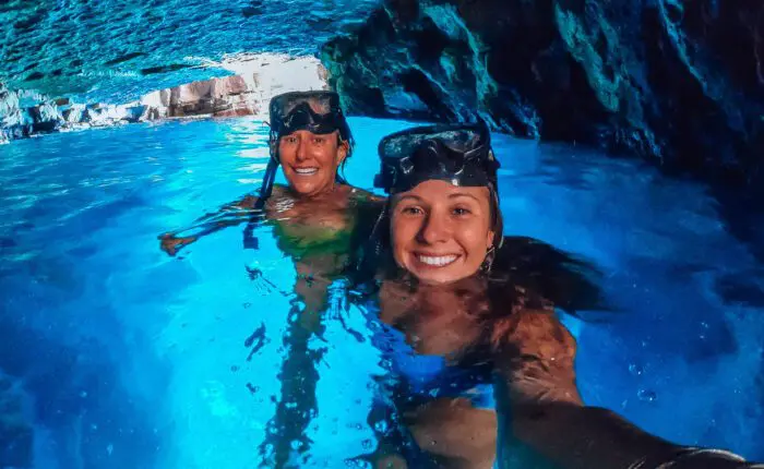 Two people wearing snorkeling gear smile in an illuminated blue cave pool, partially submerged in the vibrant water with rocky walls in the background, enjoying one of Dubrovnik's exclusive private boat tours.