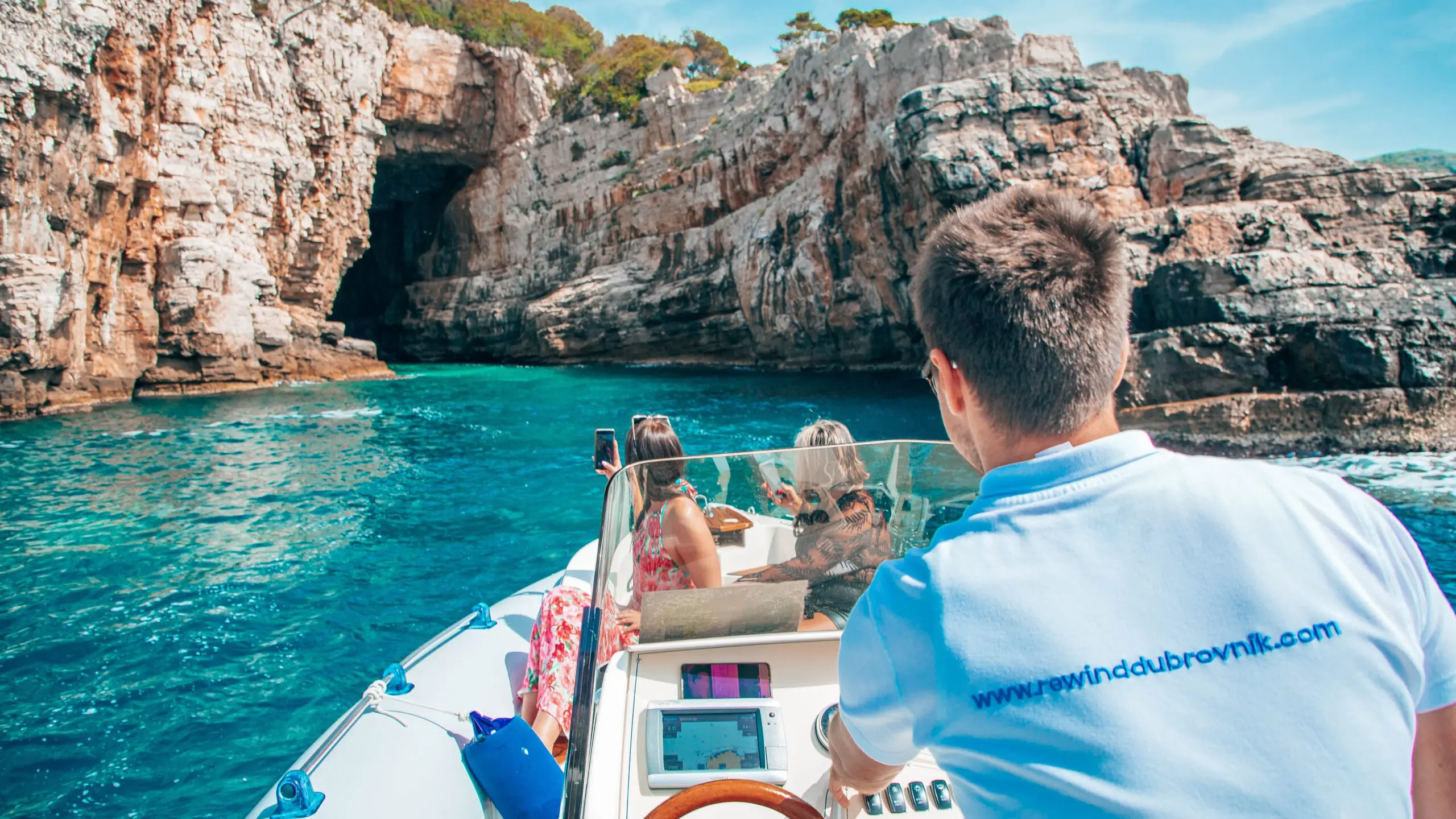 A person steers a boat towards a rocky cave as three passengers, two standing and one sitting, look at the scenery. The boat is on clear turquoise water surrounded by rocky cliffs, perfect for unforgettable private boat tours during day trips in Dubrovnik.