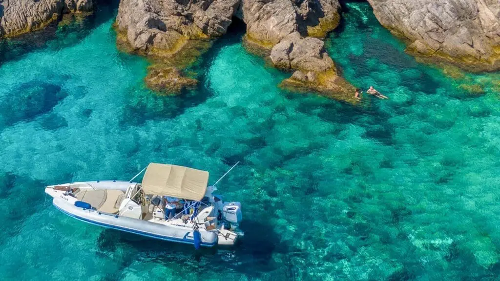 A small motorboat with a canopy floats on clear turquoise water near rocky cliffs in Dubrovnik, with a person swimming nearby, perfect for private boat tours and day trips.