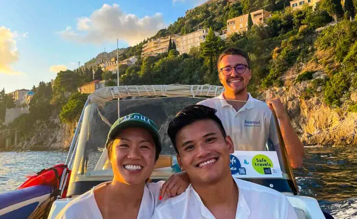 Three people are on a boat, with two individuals seated in front and one standing behind them, smiling. The backdrop consists of Dubrovnik's hillside with buildings and trees under a clear sky, creating the perfect setting for private boat tours and memorable day trips.