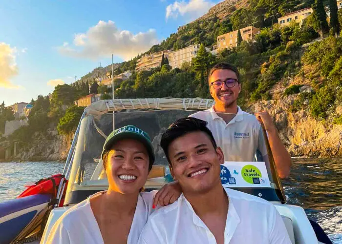 Three people are on a boat, with two individuals seated in front and one standing behind them, smiling. The backdrop consists of Dubrovnik's hillside with buildings and trees under a clear sky, creating the perfect setting for private boat tours and memorable day trips.