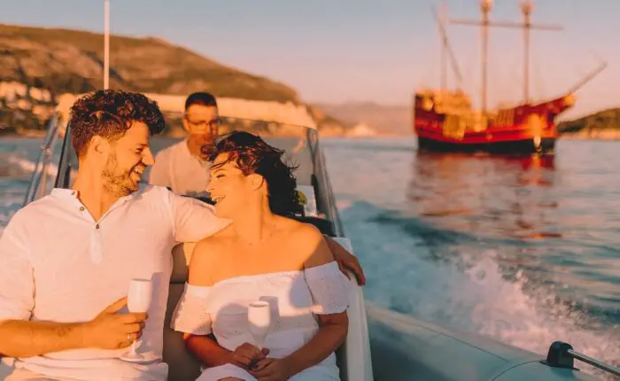 A couple holding drinks smiles on a private boat tour in Dubrovnik, with another person behind them and a large sailboat in the background on a sunny day.