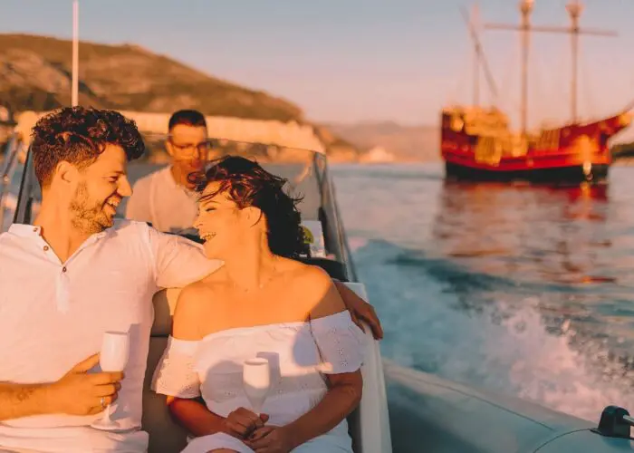 A couple holding drinks smiles on a private boat tour in Dubrovnik, with another person behind them and a large sailboat in the background on a sunny day.