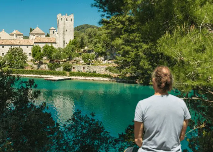 A person in a gray shirt is standing by a body of turquoise water, looking towards a historic stone building and tower surrounded by greenery, perhaps contemplating the charm of Dubrovnik's landscape on one of their private boat tours.