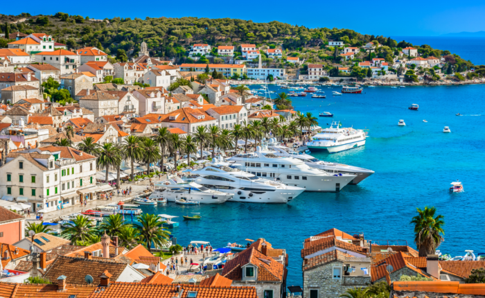 A seaside town with red-roofed buildings, palm trees, and several yachts docked in a vibrant blue bay under a clear sky. Hills and more buildings are in the background, perfect for day trips or private boat tours to explore the stunning surroundings of Dubrovnik.