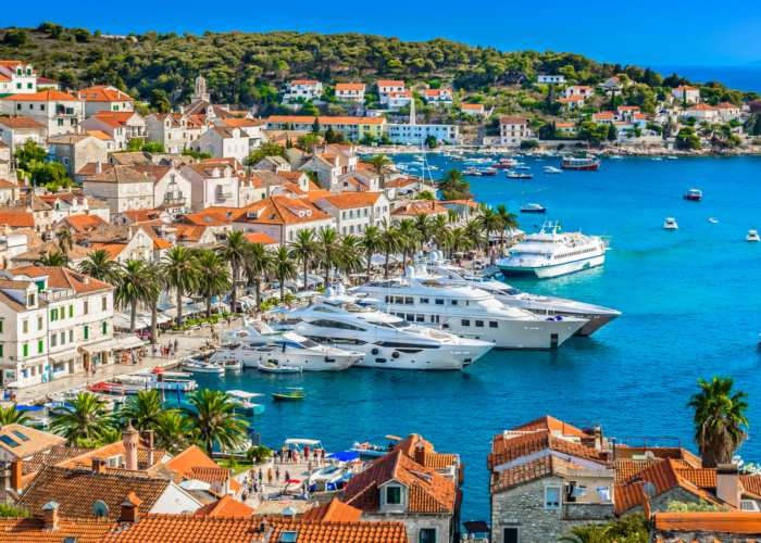 A seaside town with red-roofed buildings, palm trees, and several yachts docked in a vibrant blue bay under a clear sky. Hills and more buildings are in the background, perfect for day trips or private boat tours to explore the stunning surroundings of Dubrovnik.