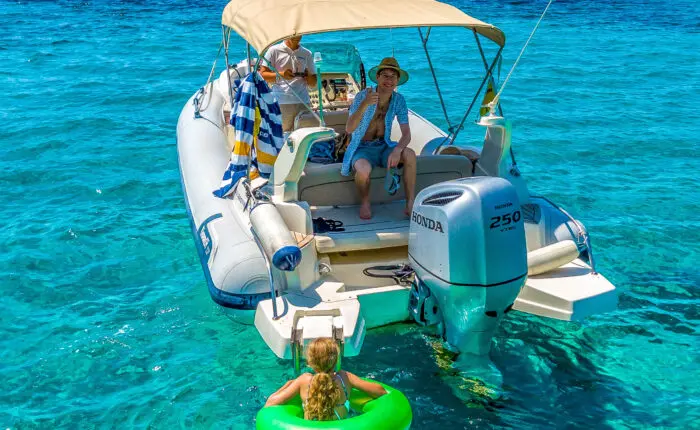 A family enjoys a sunny day on a motorboat in Dubrovnik; a child in a green float splashes in the clear blue water while an adult in a hat waves from the boat. Perfect for those seeking memorable private boat tours or day trips.