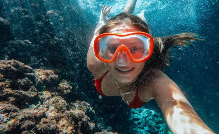 A person wearing red swim goggles and a matching red swimsuit is underwater, swimming near a coral reef off the coast of Dubrovnik. They are smiling and taking a selfie while enjoying one of their unforgettable private boat tours.