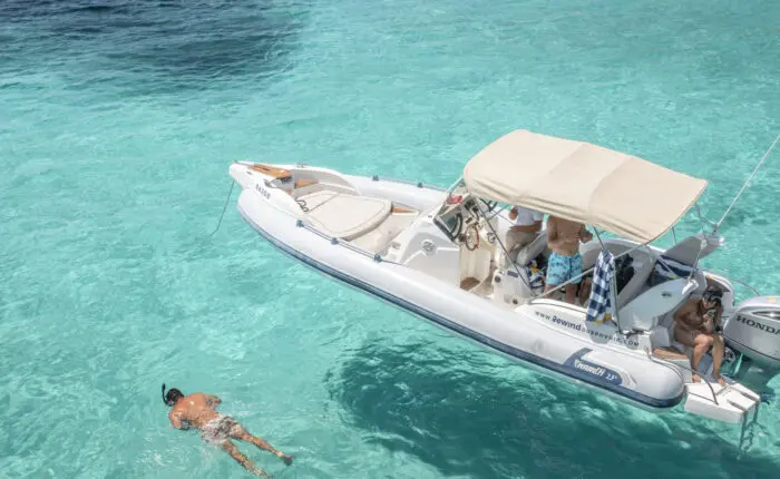 A person swims near a small motorboat on clear turquoise water, part of one of Dubrovnik's private boat tours. Several others stay on the boat, with one using binoculars. The vessel is equipped with a Honda outboard motor and a sunshade.