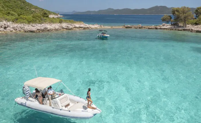 A white motorboat with a canopy floats on clear turquoise water near a rocky shore with greenery, offering an idyllic setting for private boat tours in Dubrovnik. Another boat can be seen in the background, and people are relaxing on the first boat, enjoying their day trip.