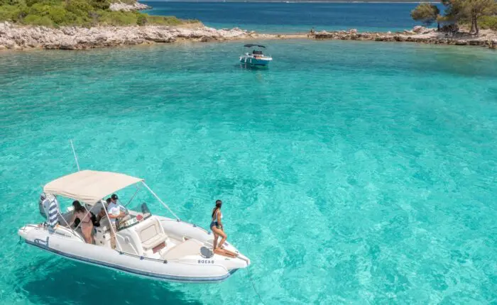 A small boat with several people onboard is anchored in clear, turquoise water near a rocky shoreline, perfect for private boat tours. Another boat is visible in the background, capturing the essence of a relaxing day trip in Dubrovnik.