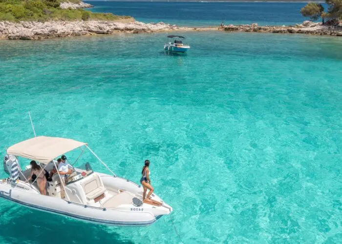 A small boat with several people onboard is anchored in clear, turquoise water near a rocky shoreline, perfect for private boat tours. Another boat is visible in the background, capturing the essence of a relaxing day trip in Dubrovnik.