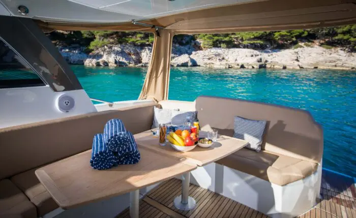 A boat's dining area, perfect for Private Boat Tours, has a table set for breakfast with fruits, juice, and rolls. Anchored near a rocky shoreline with the clear blue water of Dubrovnik and trees in the background, it's an idyllic spot for day trips.