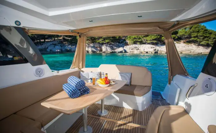 Interior of a boat with beige seating and a wooden table set with fruit and condiments, against a background of clear blue water and rocky shoreline with trees—perfect for private boat tours in Dubrovnik.