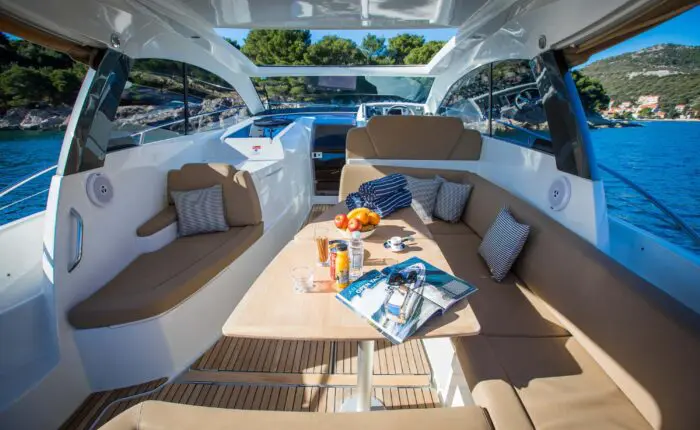 Interior view of a yacht with cushioned seating, a wooden table, and various items such as magazines, fruit, snacks, and sunglasses. The yacht is on the water near Dubrovnik with a scenic island in the background—a perfect setting for private boat tours or day trips.