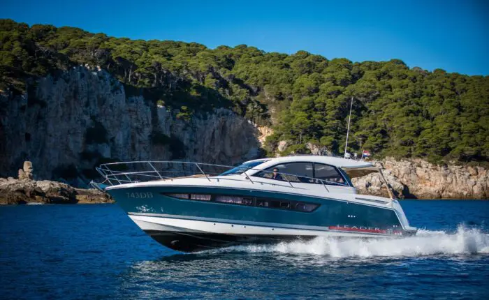 A blue and white yacht cruises through the water near a rocky, forested shoreline under a clear blue sky, perfect for private boat tours in Dubrovnik.