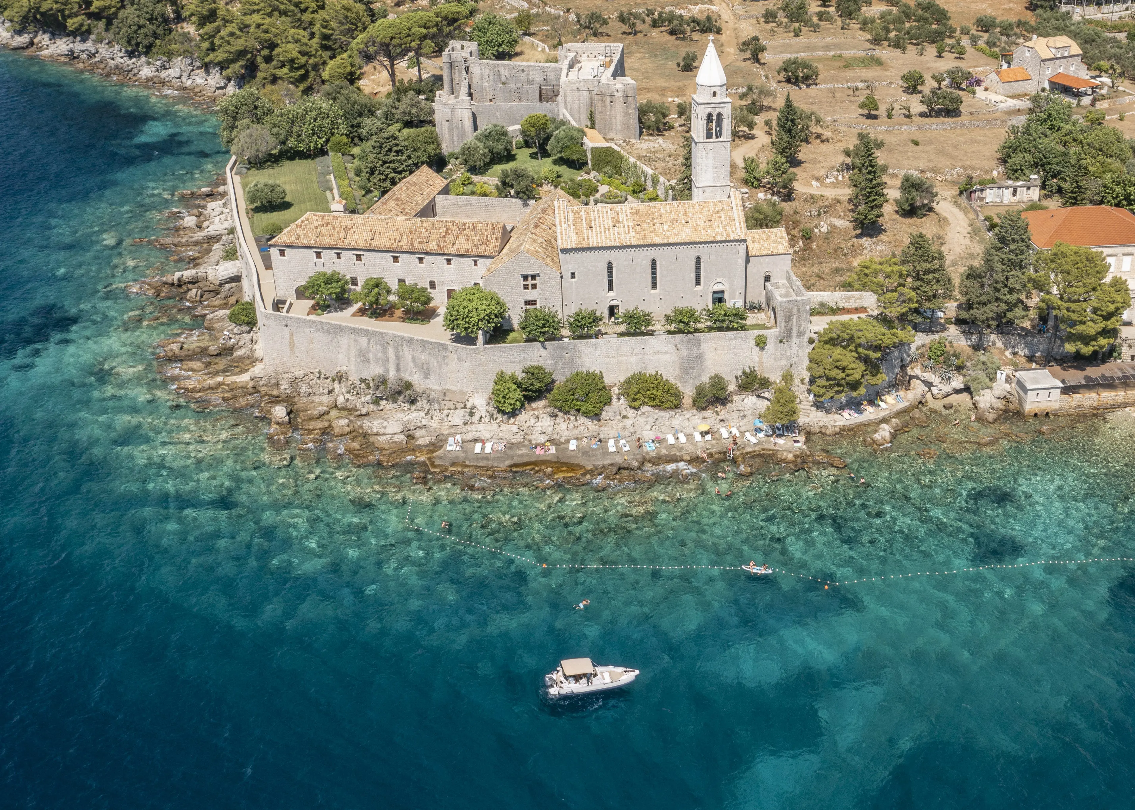 Aerial view of a historic seaside monastery in Dubrovnik with a bell tower surrounded by clear turquoise waters, a boat docked nearby, and swimming areas delineated by buoys in the foreground, making it an ideal spot for day trips or private boat tours.