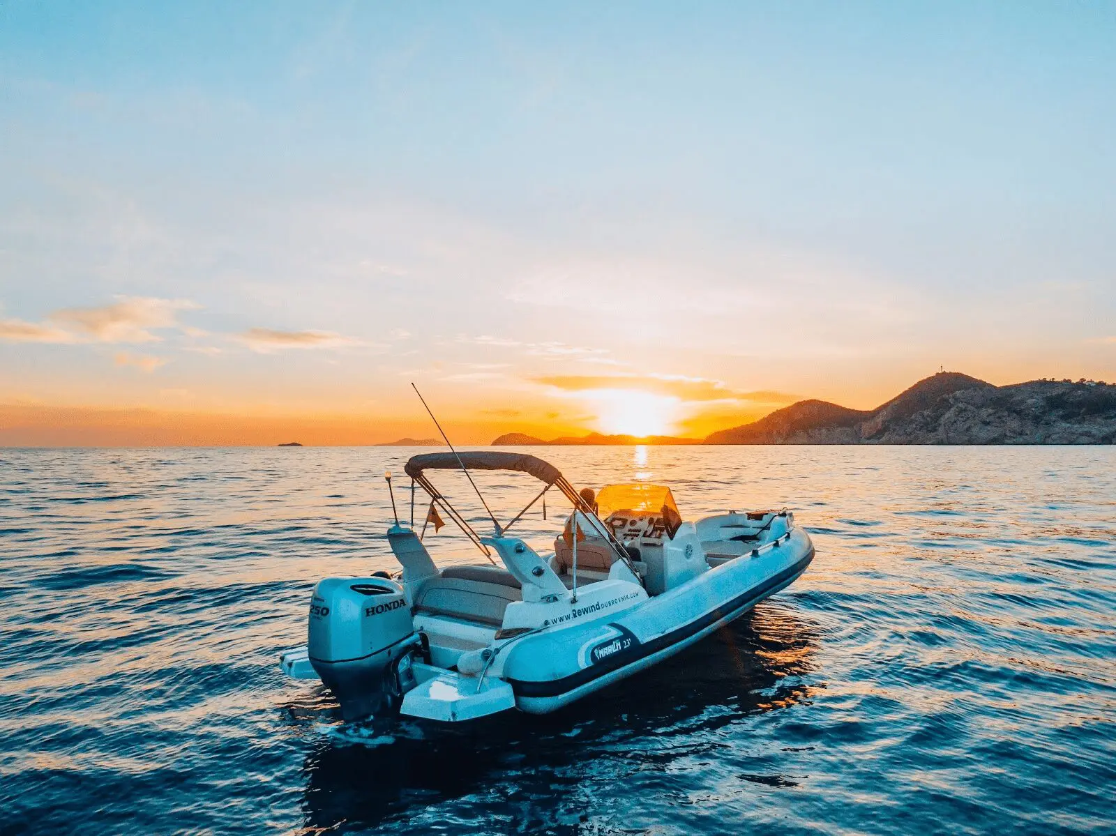 A private boat tour floats on calm water at sunset, with an orange and blue sky and silhouettes of distant mountains in the background, offering an unforgettable day trip experience near Dubrovnik.