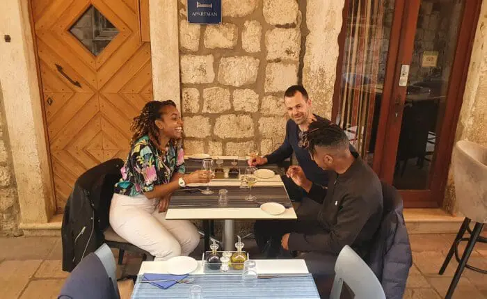 Three people seated at an outdoor restaurant table, eating and conversing, possibly discussing their recent day trips. The setting features a stone wall and wooden door in the background, reminiscent of Dubrovnik's charming architecture.