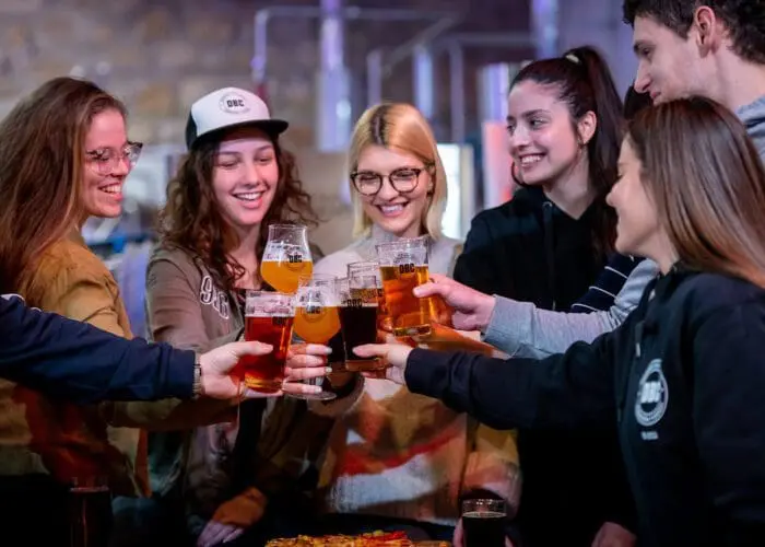 A group of seven people toasting with beer glasses, smiling and standing close together in a casual indoor setting, reminiscing about their unforgettable day trip to Dubrovnik.