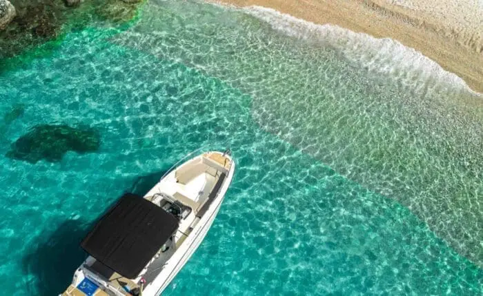 A boat with a black canopy is anchored in clear turquoise waters near a rocky shoreline with a sandy beach, perfect for day trips or private boat tours around Dubrovnik.