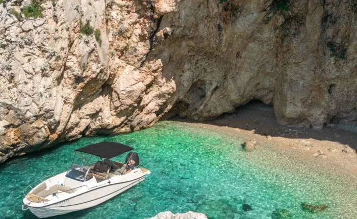 A small white boat with a black canopy is anchored in clear turquoise water near a rocky cliff and a small beach with large rocks, perfect for Private Boat Tours in Dubrovnik.