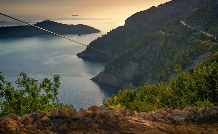 A coastal landscape at sunset near Dubrovnik, featuring hills with lush greenery, a winding road perfect for day trips, and islands in the distance. Cable lines run through the tranquil scene above calm waters, ideal for private boat tours.