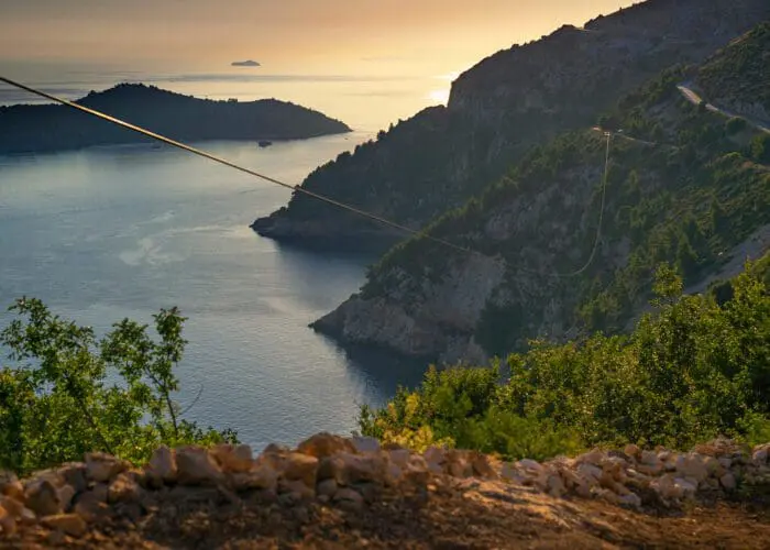 A coastal landscape at sunset near Dubrovnik, featuring hills with lush greenery, a winding road perfect for day trips, and islands in the distance. Cable lines run through the tranquil scene above calm waters, ideal for private boat tours.