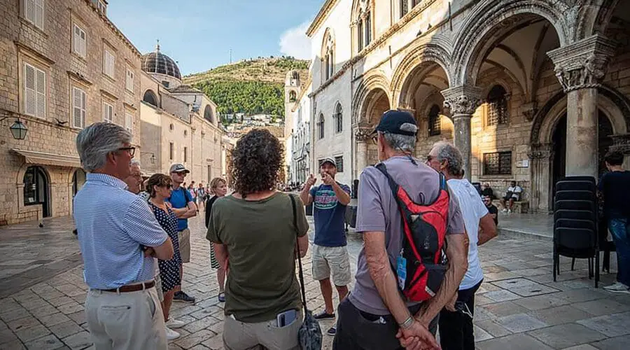 A group of tourists listens to a guide in Dubrovnik's old town square, surrounded by historic architecture, under a sunny sky. The guide mentions how private boat tours and day trips offer unique perspectives on the city's charming coastline.