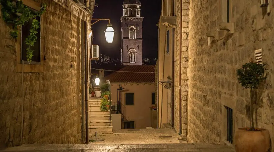 Narrow stone street at night with illuminated church bell tower in the background, typical of Dubrovnik's charm. Potted plant and hanging lantern visible along the textured walls, making it a perfect spot for evening strolls after day trips.