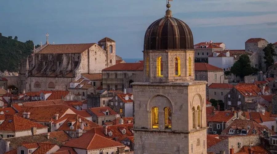 A scenic view of Dubrovnik's historic city with red-tiled rooftops and a central bell tower illuminated at dusk invites you to explore further. Enhance your visit by enjoying exclusive Private Boat Tours or unforgettable Day Trips.