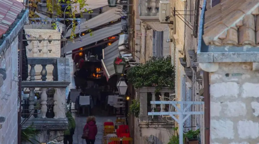 A narrow cobblestone street lined with buildings and outdoor seating. Pedestrians walk toward cafes sheltered by awnings. Balconies and plants decorate the facades of the buildings, creating a picturesque setting reminiscent of charming day trips in Dubrovnik.