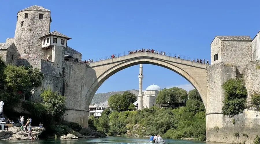 A stone arch bridge spans a river, with people walking on it. Historic buildings and greenery surround the area under a clear blue sky. A small boat offers private boat tours on the river below the bridge, making it an ideal spot for day trips in Dubrovnik.