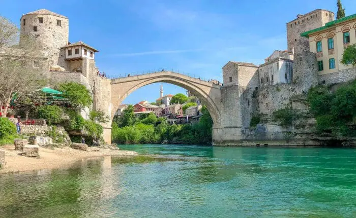 A stone arch bridge spans a river between historical buildings on both sides, under a clear blue sky with green water and lush vegetation along the riverbanks, much like the picturesque scenes you would encounter on Dubrovnik day trips or private boat tours.