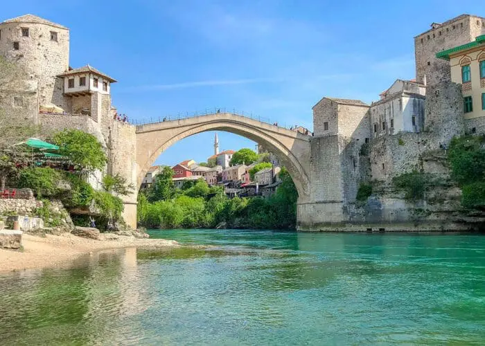 A stone arch bridge spans a river between historical buildings on both sides, under a clear blue sky with green water and lush vegetation along the riverbanks, much like the picturesque scenes you would encounter on Dubrovnik day trips or private boat tours.