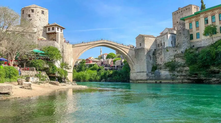 A stone arch bridge spans a river between historical buildings on both sides, under a clear blue sky with green water and lush vegetation along the riverbanks, much like the picturesque scenes you would encounter on Dubrovnik day trips or private boat tours.