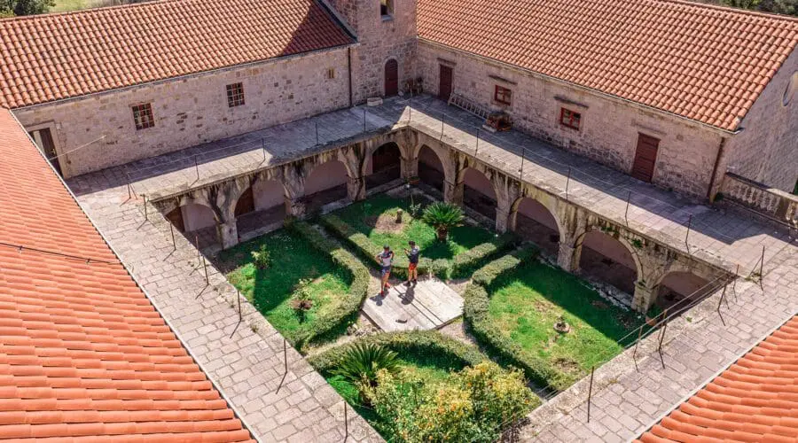 Aerial view of a courtyard garden surrounded by a building with red-tiled roofs and arches in Dubrovnik. Two people stand on a walkway in the center of the garden. Perfect for day trips or private boat tours nearby, this spot captures the city’s enchanting beauty.