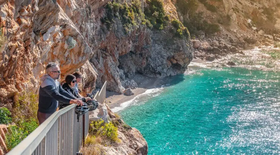 Two people stand at a railing on a rocky cliff overlooking the clear blue sea near Dubrovnik, with sunlight reflecting on the water; lush greenery and a small beach are visible below, making it an ideal spot for day trips or private boat tours.