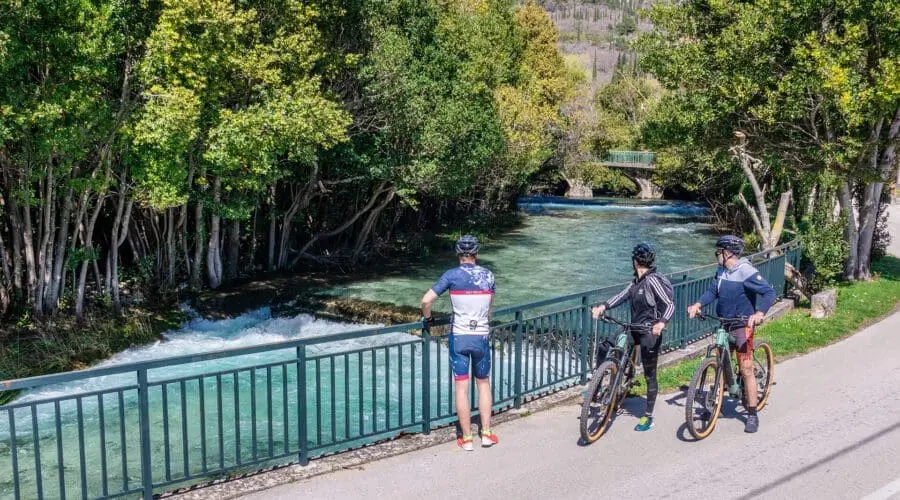 Three cyclists pause on a riverside path to appreciate the scenic view of a flowing river surrounded by lush greenery, reminiscent of the picturesque waters seen during private boat tours in Dubrovnik.