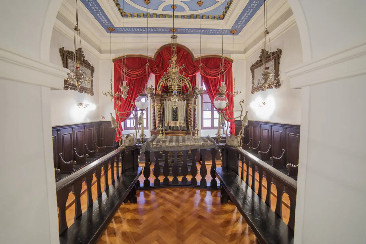 The interior of this historic synagogue, a highlight on any Jewish Tour in Dubrovnik, showcases ornate woodwork, a central platform adorned with red curtains, decorative hanging lamps, and an intricately designed ceiling that speaks volumes of the enduring Jewish Legacy.