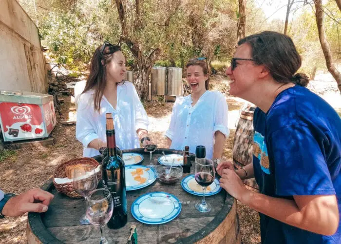 Three people enjoy a casual outdoor meal around a rustic barrel table in Dubrovnik, sharing wine and snacks while laughing together, reminiscent of the joy found on private boat tours or day trips.