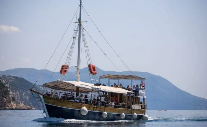 A wooden sightseeing boat with passengers sails on a calm sea during one of the popular day trips, with hilly terrain and a cloudy sky in the background. Consider Dubrovnik for memorable private boat tours amidst stunning scenery.