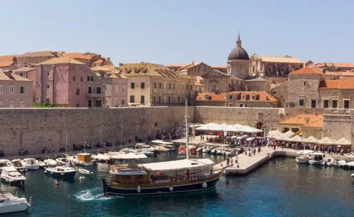 A harbor with docked boats, waterfront buildings with red-tiled roofs, and a prominent dome in the background. A large wooden boat is navigating through the water—perfect for private boat tours or day trips to explore the beauty of Dubrovnik.