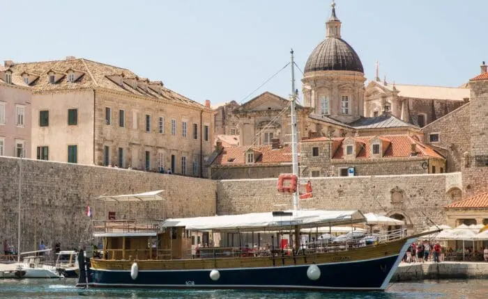 A traditional wooden boat docked at the historical port of Dubrovnik, with old stone buildings and a large domed church in the background, offers a glimpse into a bygone era. Perfect for private boat tours or day trips, this scene captures the essence of timeless maritime charm.